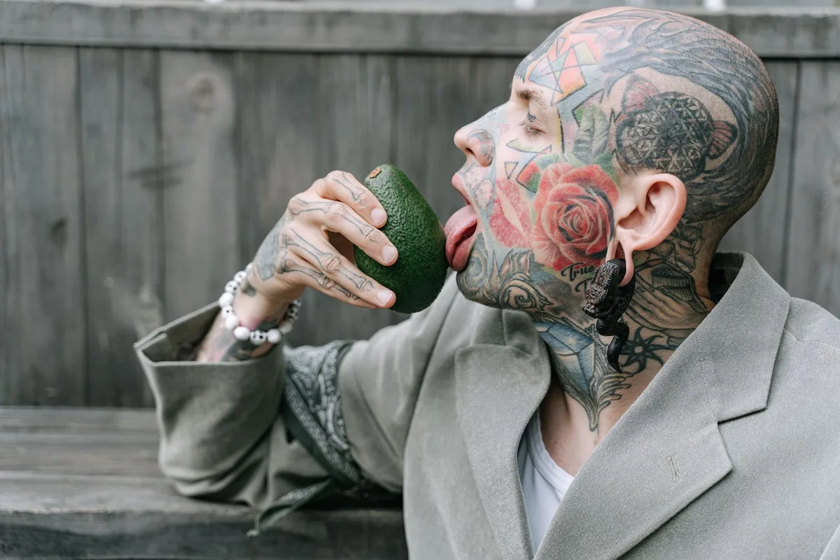 Stylish tattooed man licking an avocado in an artistic portrait setting.