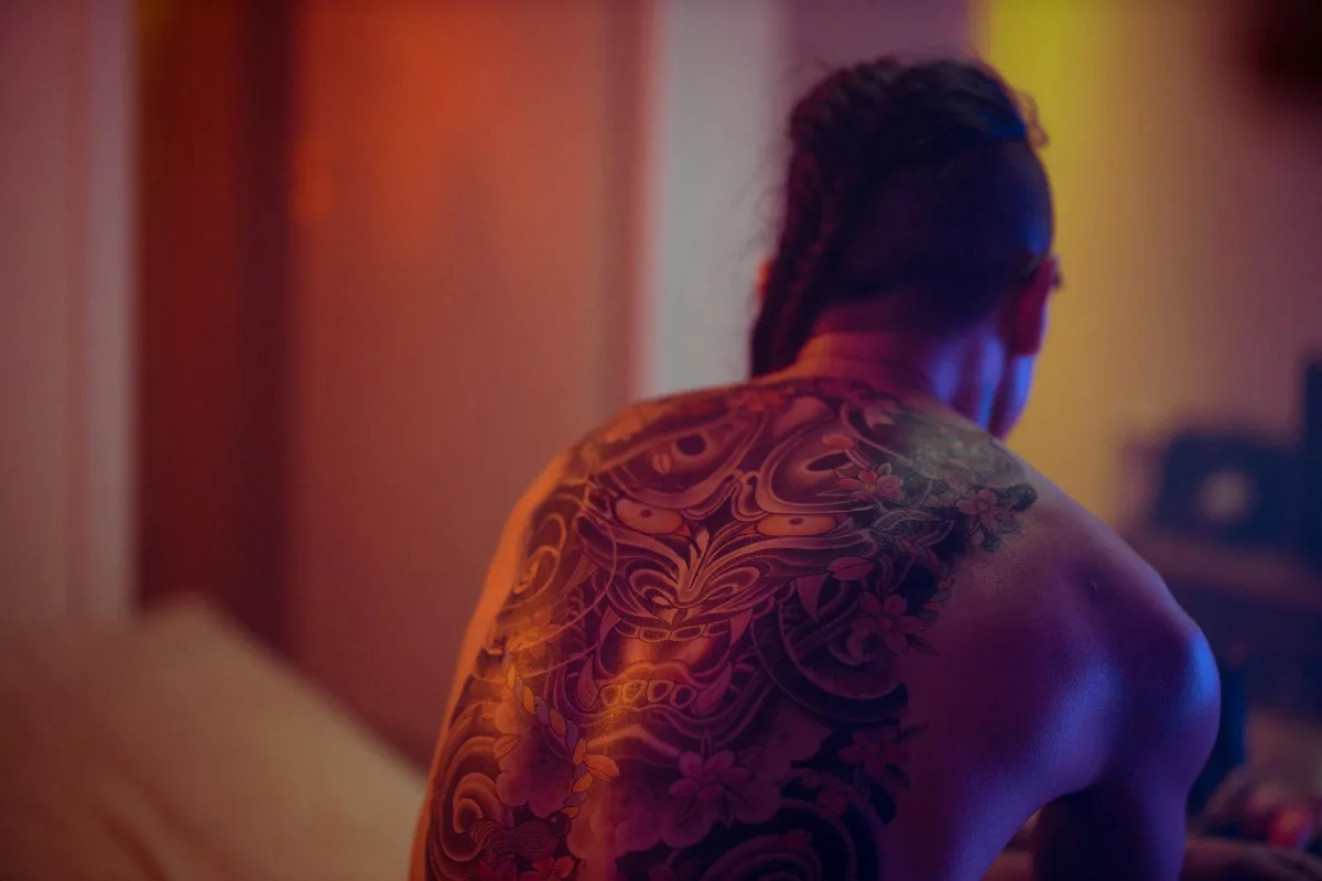 Artistic photo of a shirtless individual from behind, showcasing a detailed back tattoo in warm indoor lighting.