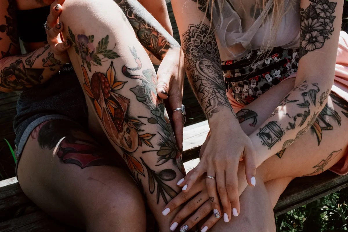 Close-up of tattooed women's legs and arms, showcasing colorful and intricate designs in an outdoor setting.