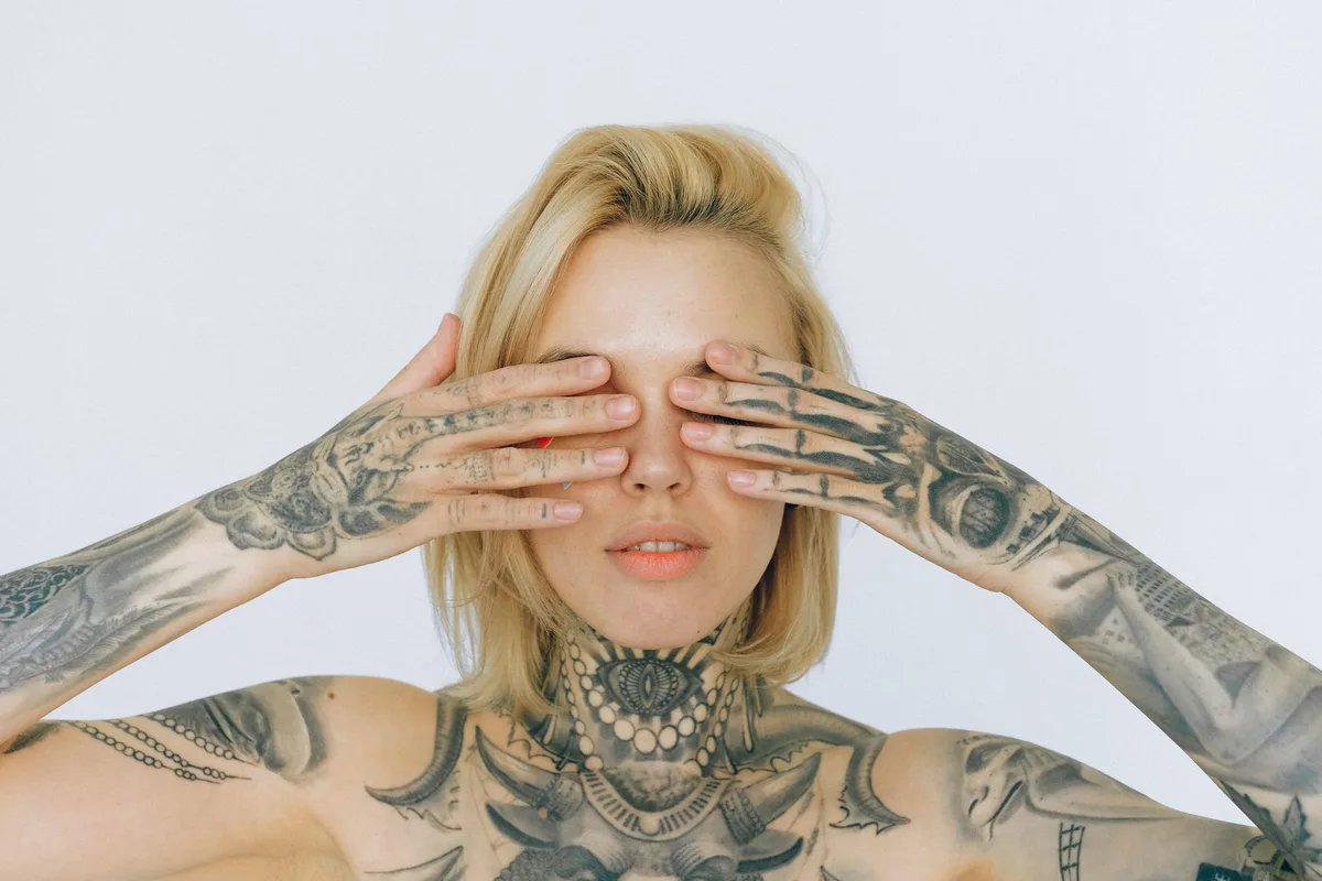Close-up image of a tattooed woman with blond hair covering her eyes with her tattooed hands.