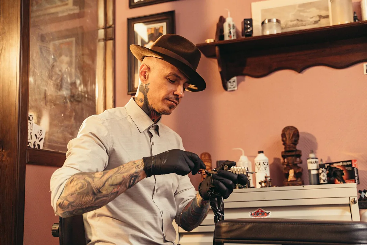 Professional tattoo artist working meticulously in his studio, wearing gloves and a fedora hat.