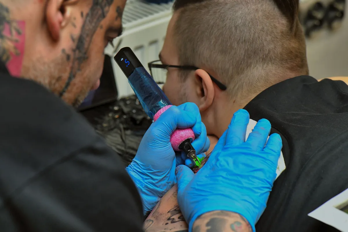 Close-up of a tattoo artist tattooing a client's shoulder with blue gloves and a tattoo machine.
