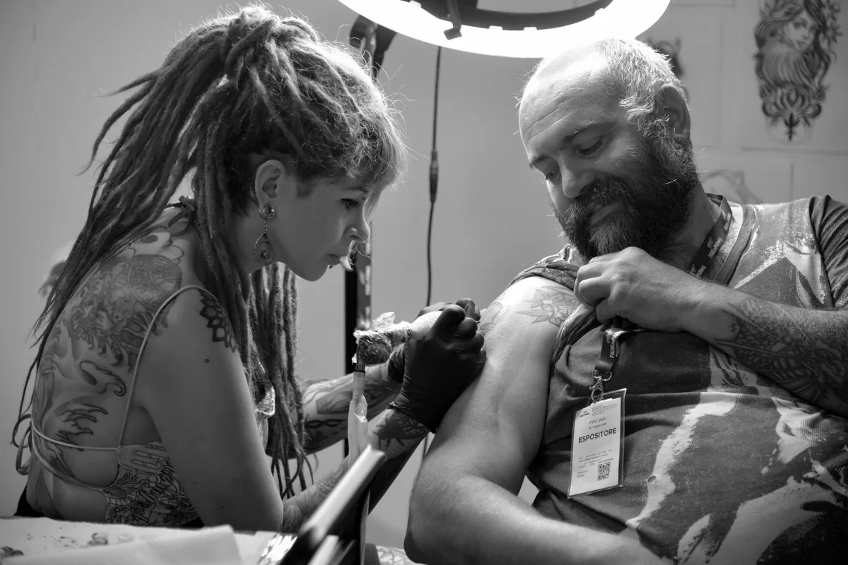 A tattoo artist with dreadlocks works on a client's arm in a studio, captured in black and white.