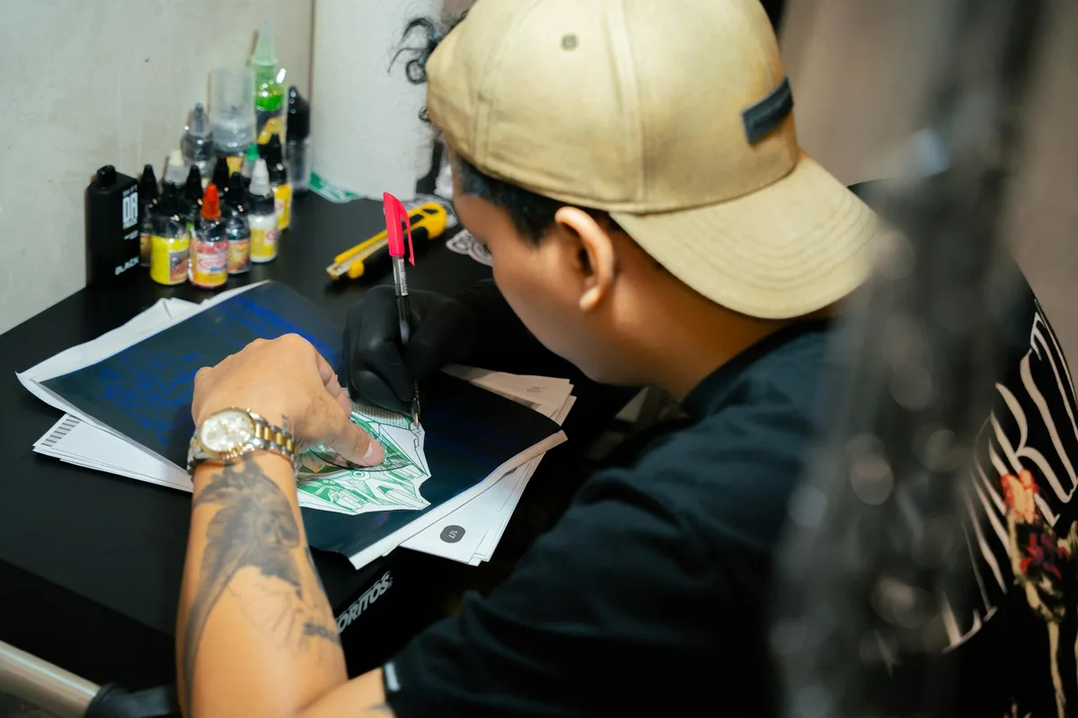 Tattoo artist drawing a new design on paper, surrounded by ink bottles in a studio.