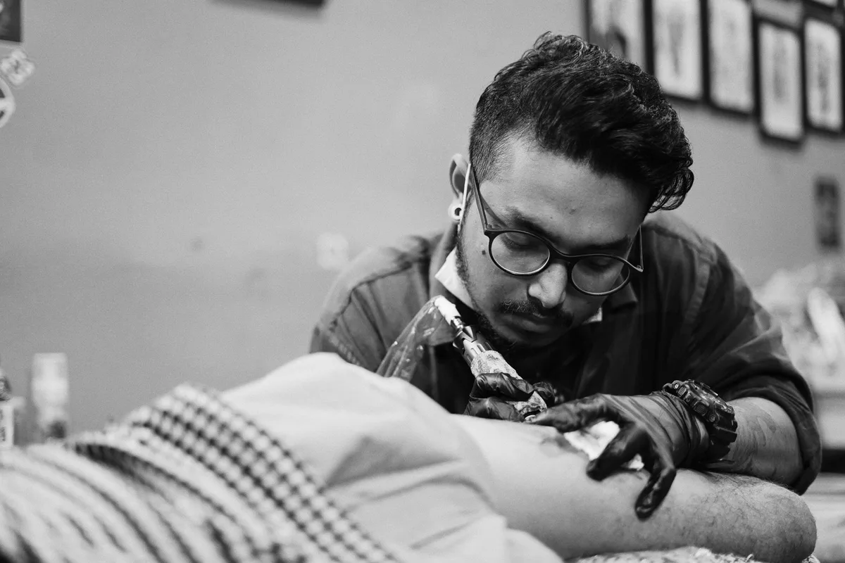 Black and white ethnic man in glasses and gloves using tattoo machine while working faceless client in salon
