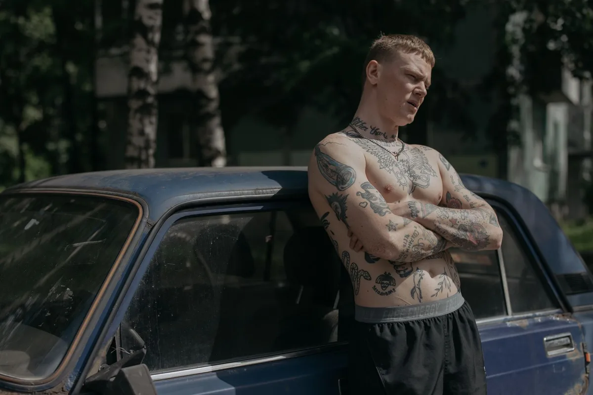 A heavily tattooed man stands shirtless, leaning on a vintage car in daylight.