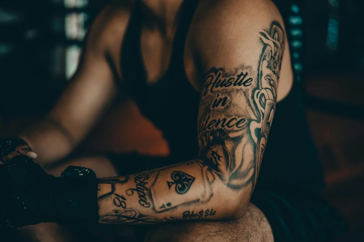 Close-up of tattooed arm with motivational text in a dimly lit setting, emphasizing strength and resilience.