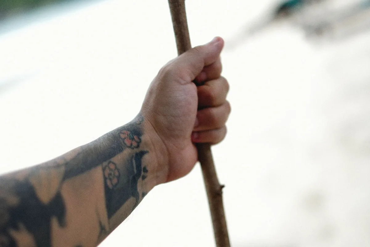 A tattooed hand firmly holds a wooden stick against a blurred natural background.