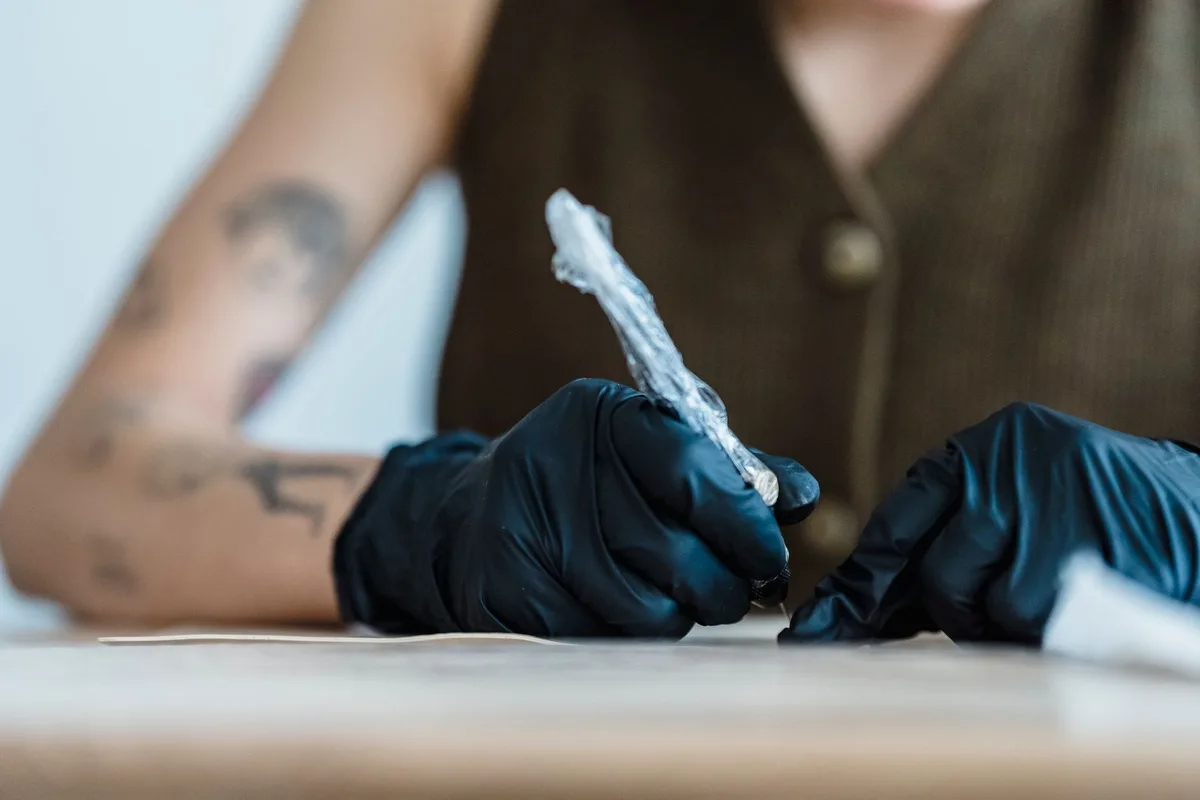 Crop anonymous artist in black gloves drawing with tattoo machine on practice skin on blurred background