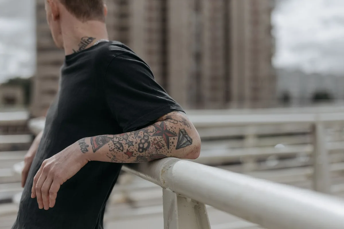 Stylish tattooed man leans on a railing in a city setting. Urban lifestyle captured outdoors.